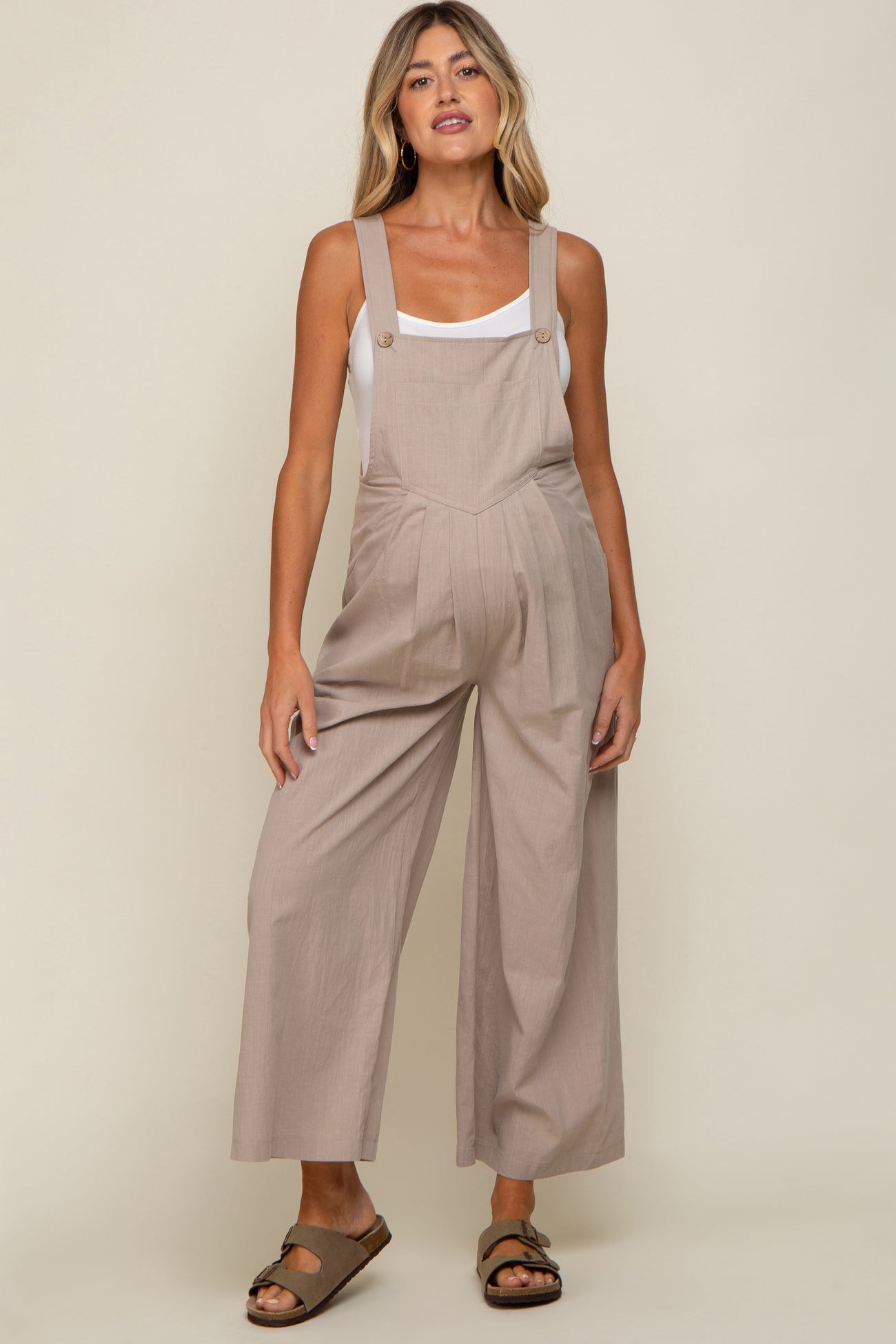woman wearing a light brown maternity jumpsuit with wide legs and buttons, showcasing her baby bump.