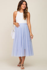woman wearing a white sleeveless top and a light blue maternity tulle skirt, holding her baby bump with a handbag