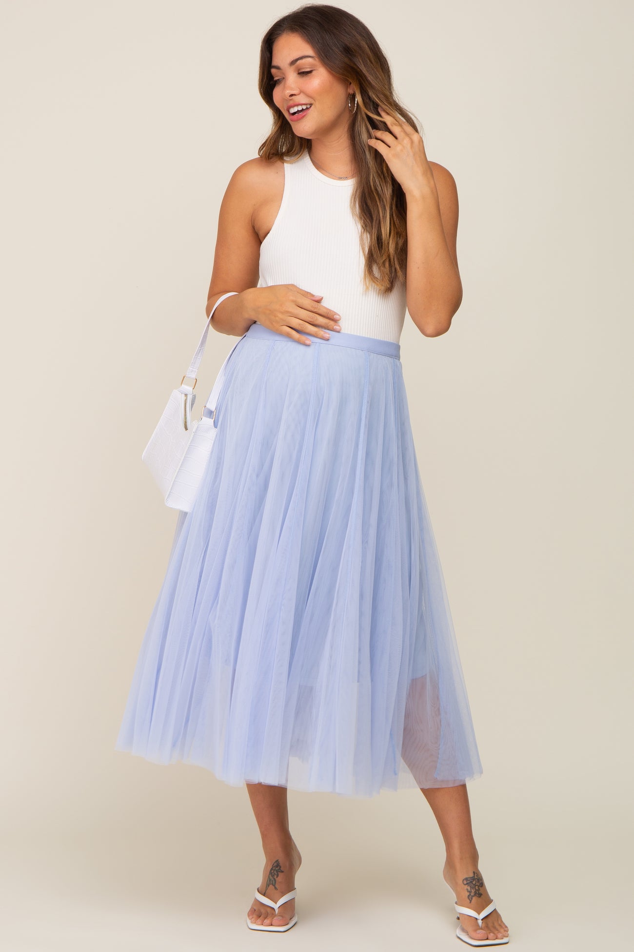Pregnant woman wearing a white ribbed tank top and light blue tulle midi skirt with a white handbag