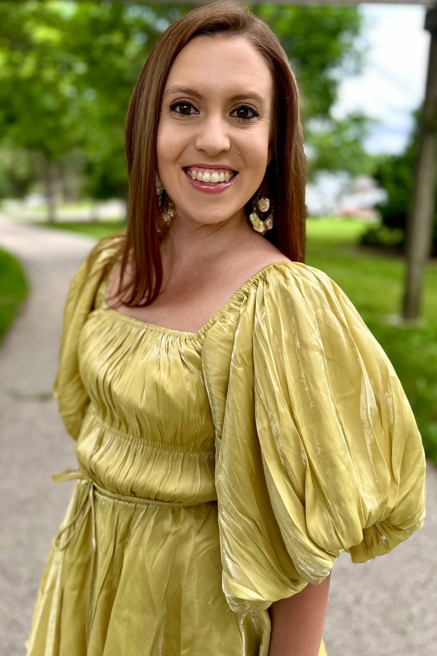 woman wearing a yellow puff-sleeve dress with a smocked bodice, smiling outdoors with greenery in the background