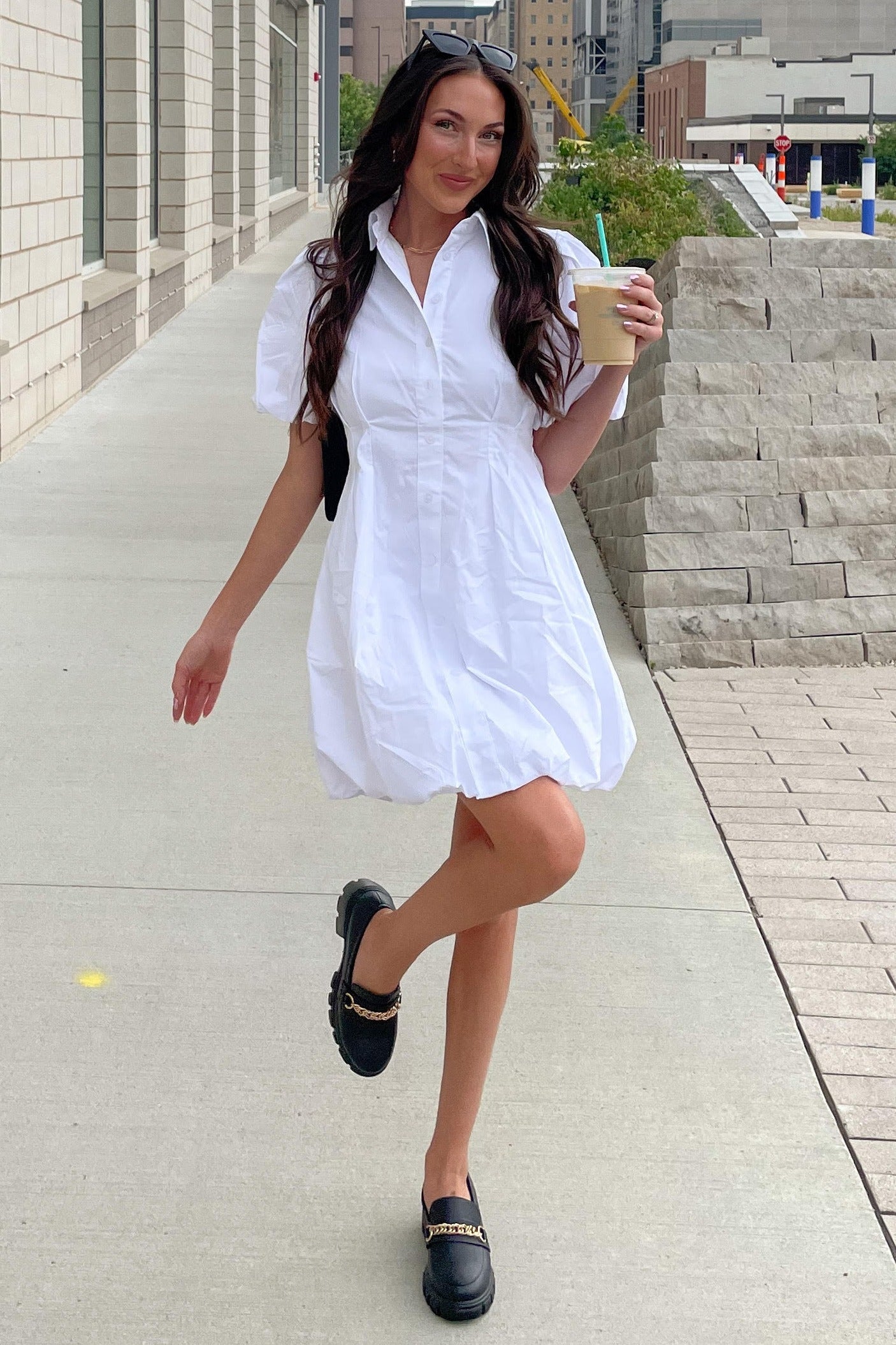 woman wearing a white short sleeve shirt dress and black clogs, holding a drink while walking outdoors