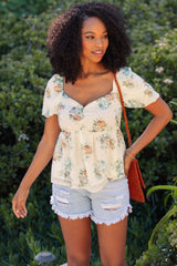 woman wearing a white floral peplum top and denim shorts, standing outdoors with a textured bag over her shoulder