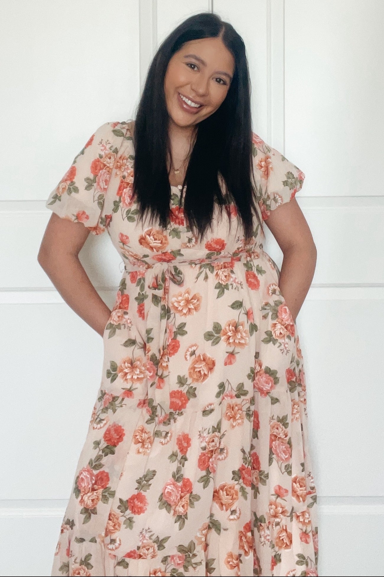 woman wearing a flowy pastel floral maxi dress with short sleeves and a tied waist, smiling and standing with hands in pockets
