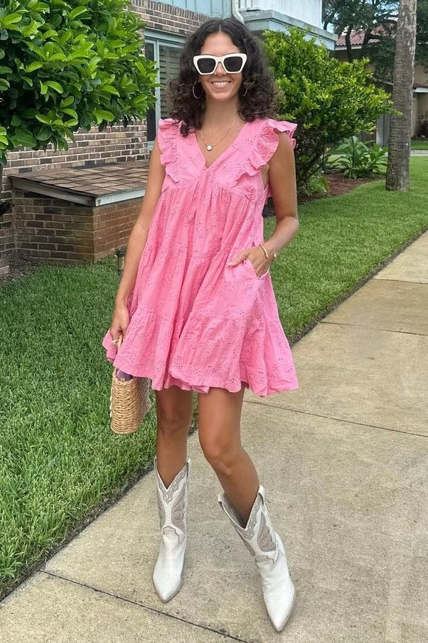 woman wearing a pink ruffled sundress and white cowboy boots, holding a straw bag, standing on a sidewalk with greenery around