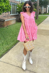 woman wearing a pink ruffled sleeveless dress, white cowboy boots, and holding a woven handbag with sunglasses on