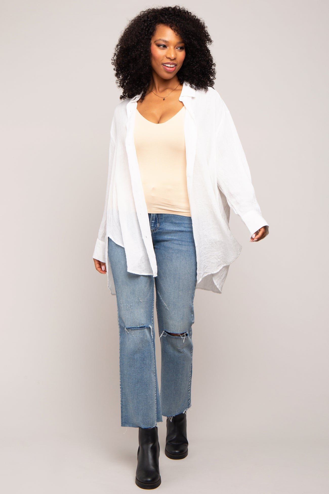 woman wearing a white oversized button-up shirt, beige tank top, and distressed blue jeans with black ankle boots