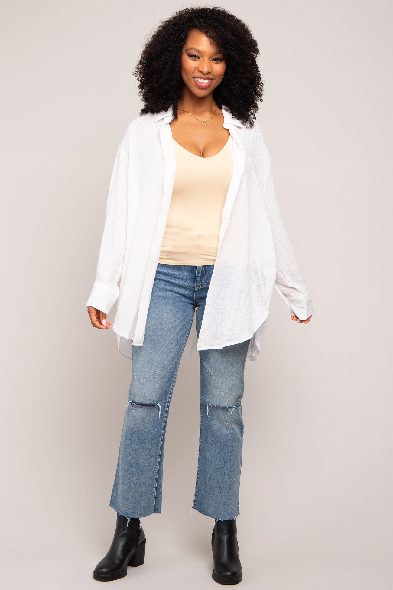 woman wearing an oversized white button-up shirt over a beige tank top and paired with distressed light blue jeans and black heeled boots