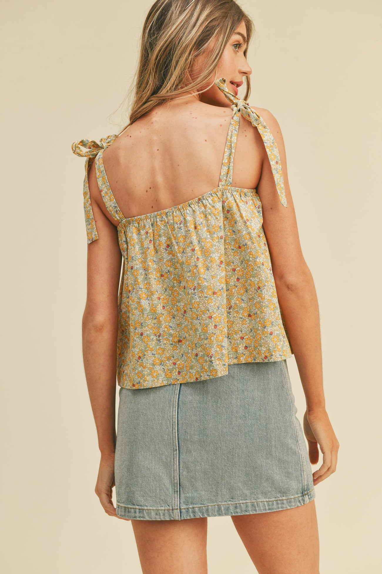 woman wearing a yellow floral print tank top with tie straps paired with a denim mini skirt, back view