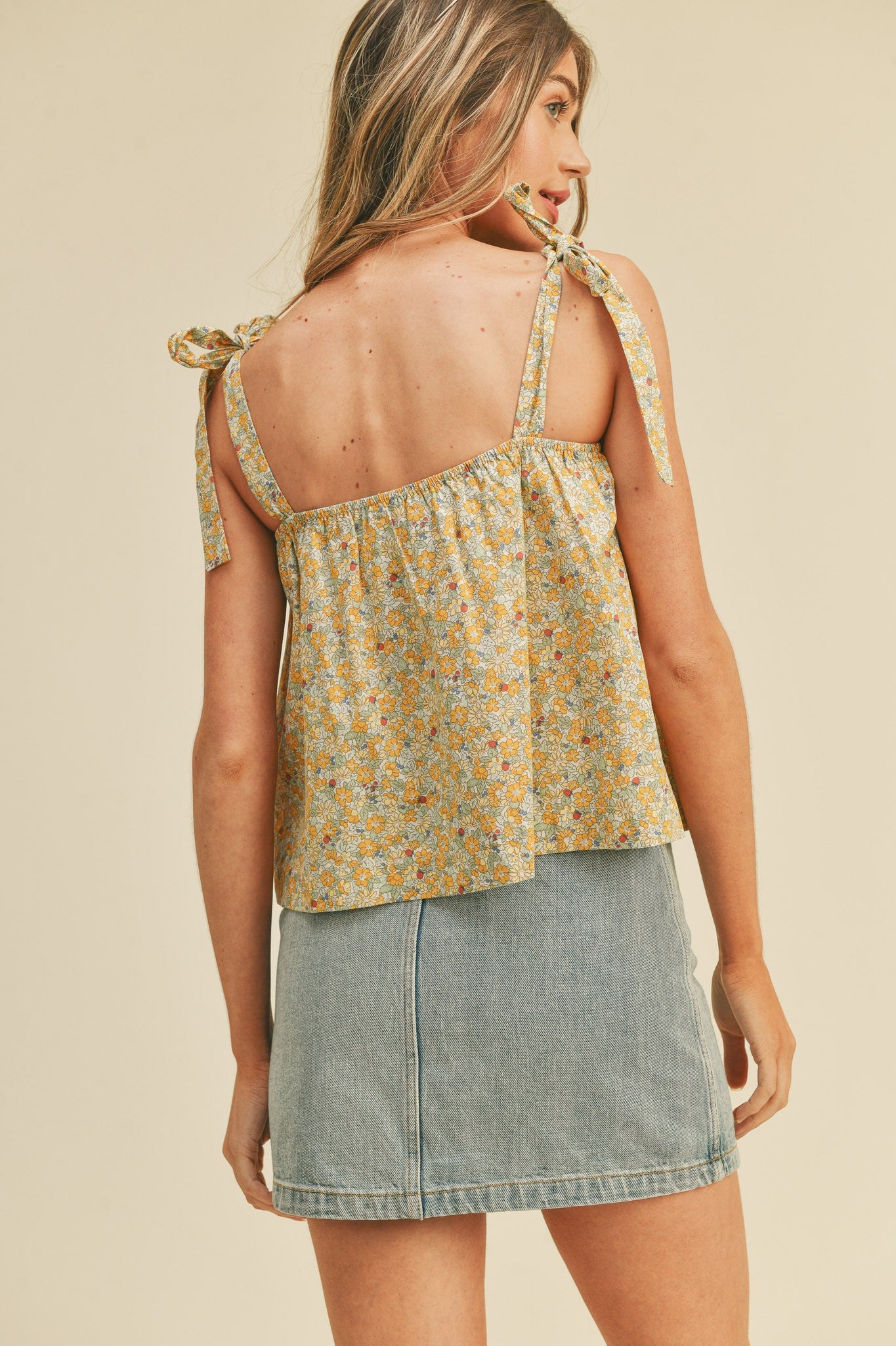 woman wearing a yellow floral print tank top with tie straps paired with a denim mini skirt, back view