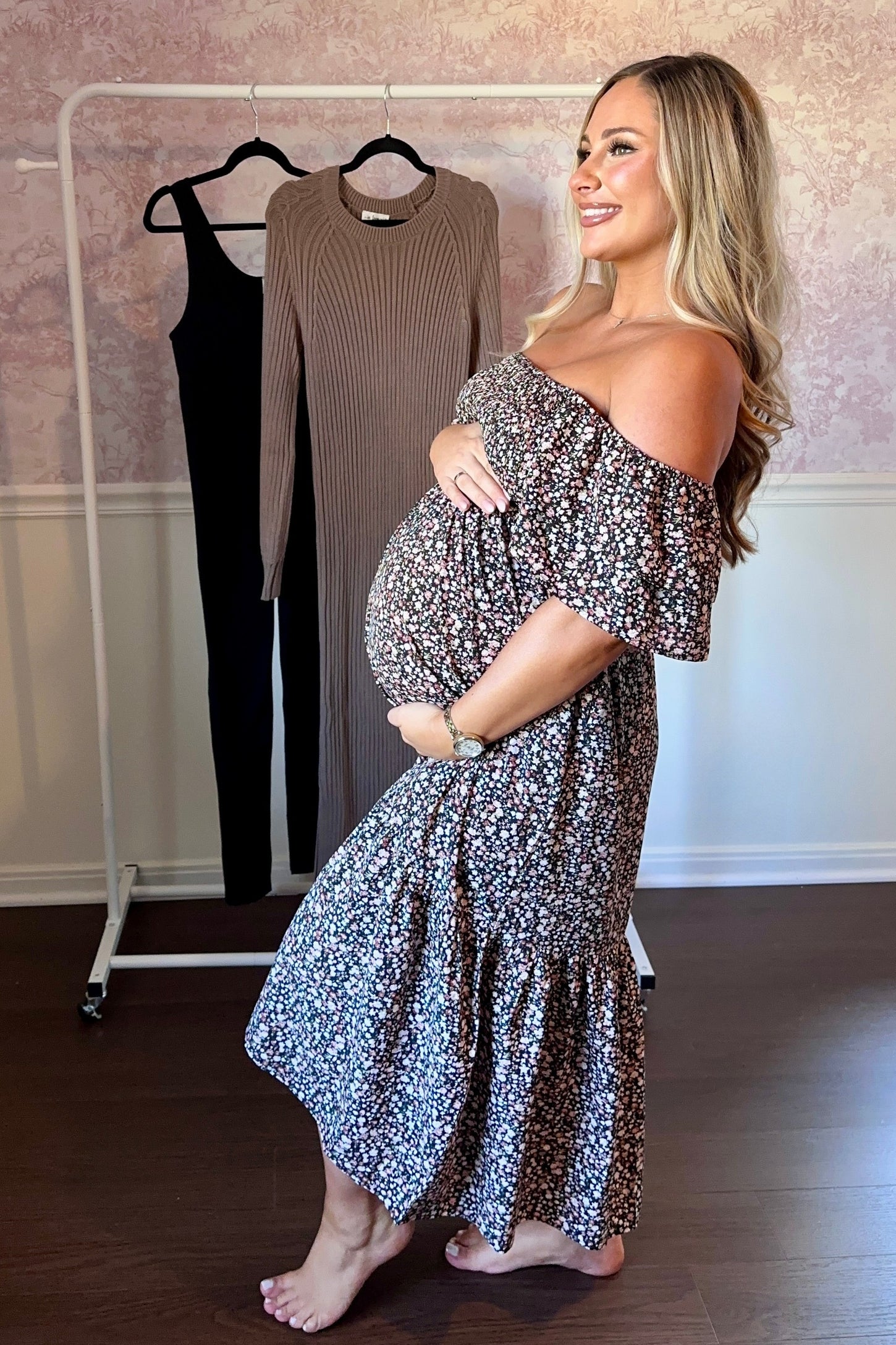 woman wearing a floral off-shoulder maternity dress and holding her baby bump, with a ribbed dress hanging in the background
