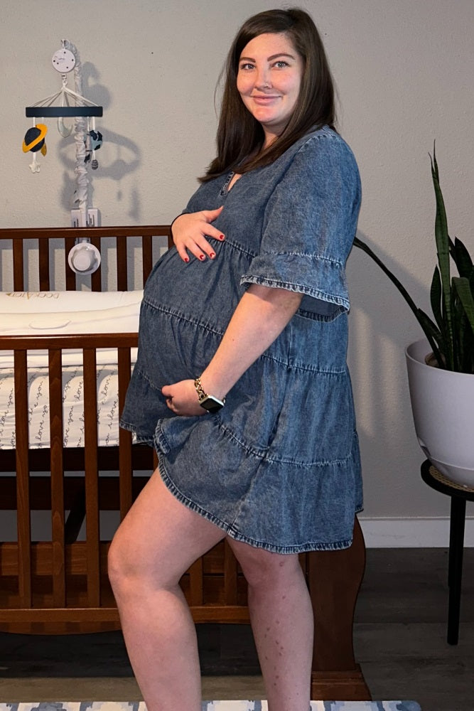 woman wearing a denim maternity tiered dress and holding her baby bump, standing next to a crib