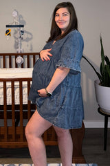 woman wearing a denim maternity tiered dress and holding her baby bump, standing next to a crib