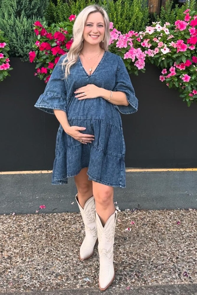 woman wearing a denim maternity dress with flared sleeves and holding her baby bump, standing in front of flowers