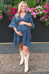 woman wearing a denim maternity dress with flared sleeves and holding her baby bump, standing in front of flowers