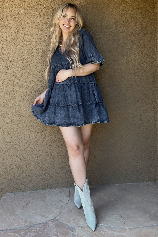 woman wearing a dark denim tiered maternity dress with short sleeves and cowboy boots, standing against a textured wall