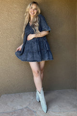 woman wearing a dark denim tiered maternity dress with short sleeves and cowboy boots, standing against a textured wall