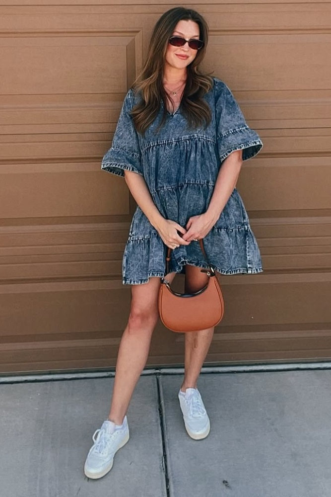 woman wearing a denim tiered dress and holding a brown handbag, posing casually with sunglasses on