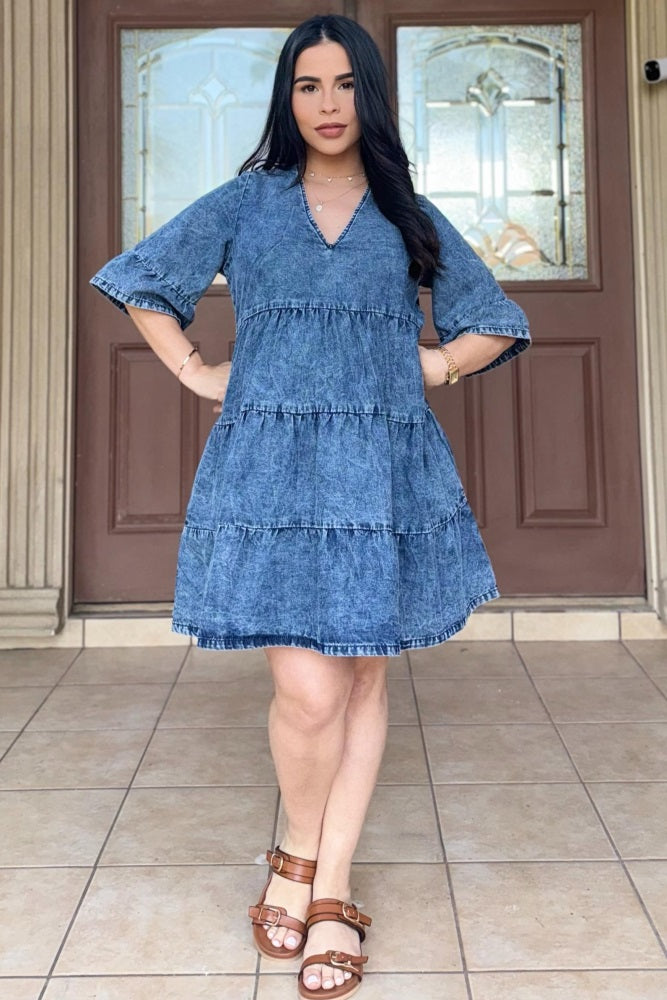 woman wearing a denim tiered maternity dress with short sleeves and a v-neckline, standing in front of a double door