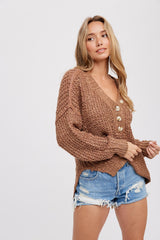 Woman wearing a brown chunky knit button-up cardigan and distressed denim shorts against a white background