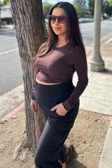 woman wearing a brown long sleeve maternity top and black maternity pants, showing her baby bump while standing outdoors