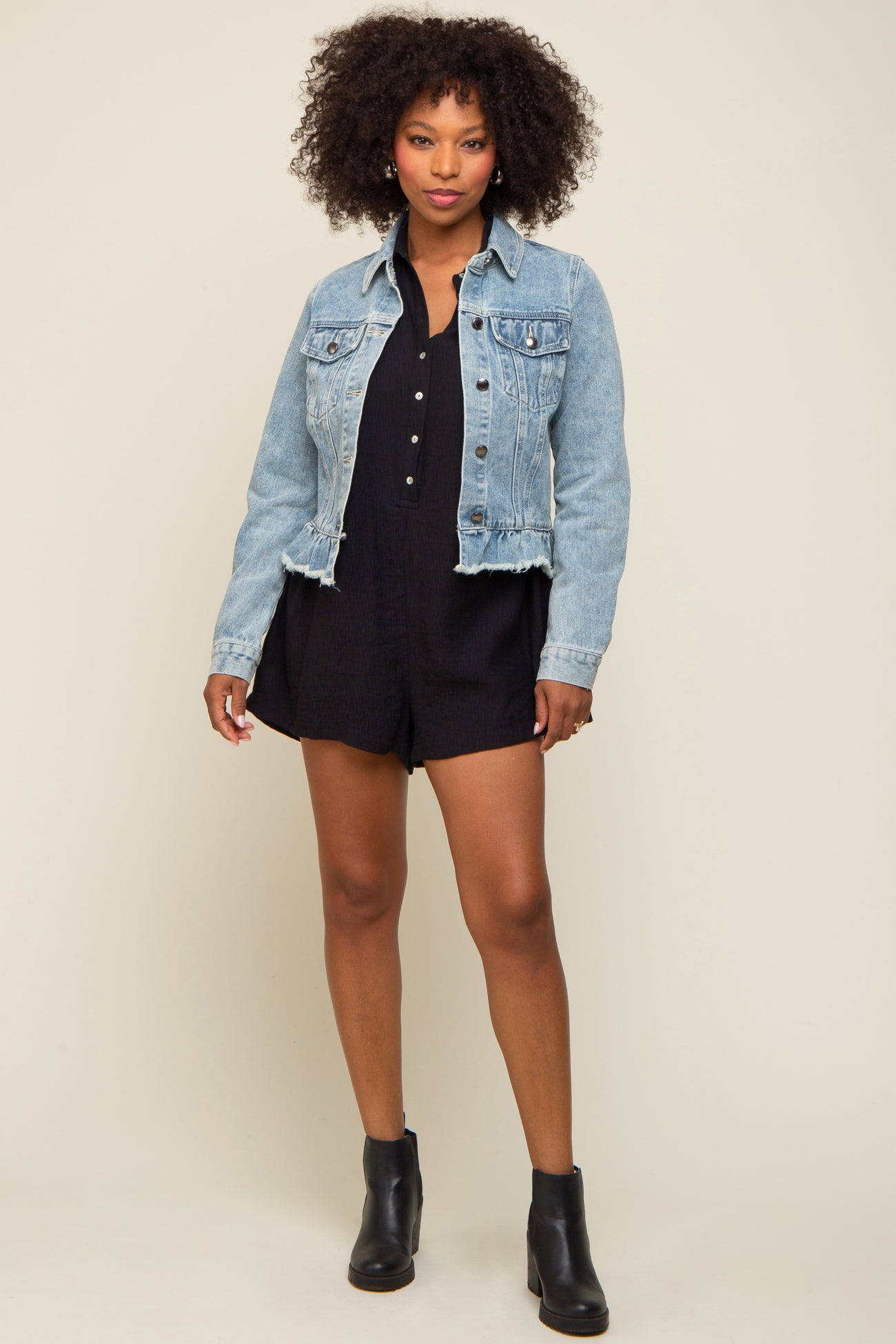 woman wearing a light wash denim jacket over a black romper with buttons and ankle boots
