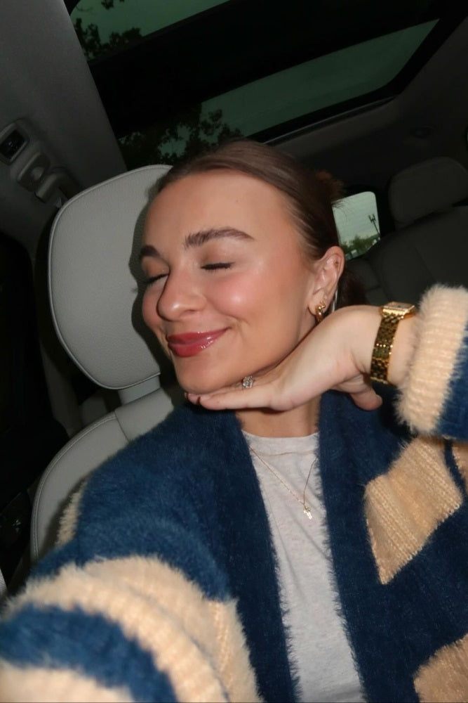 Smiling woman with eyes closed wearing a blue and tan striped fuzzy cardigan and gold watch in a car