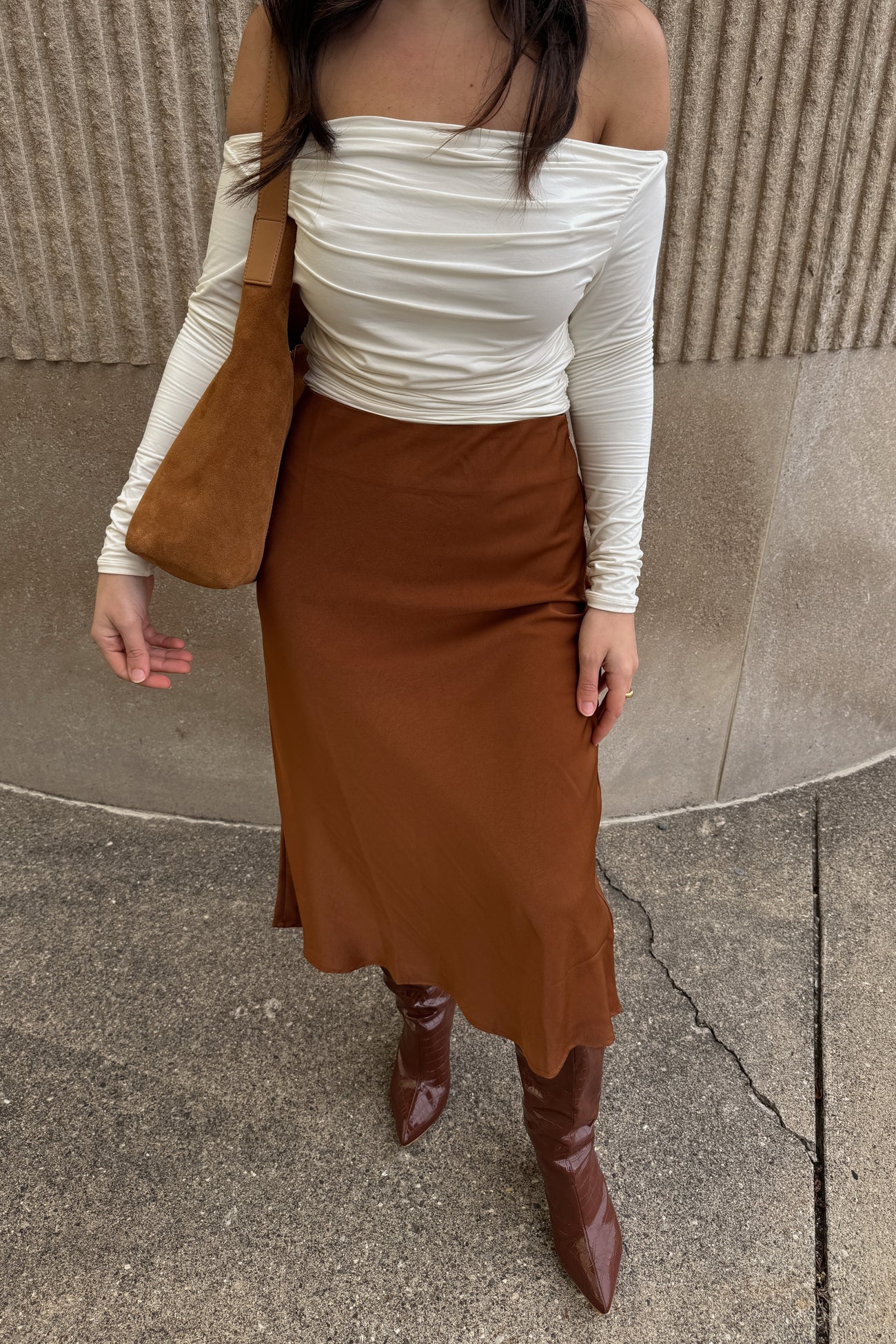 woman wearing white off-the-shoulder top, brown satin midi skirt, and brown leather boots with suede bag