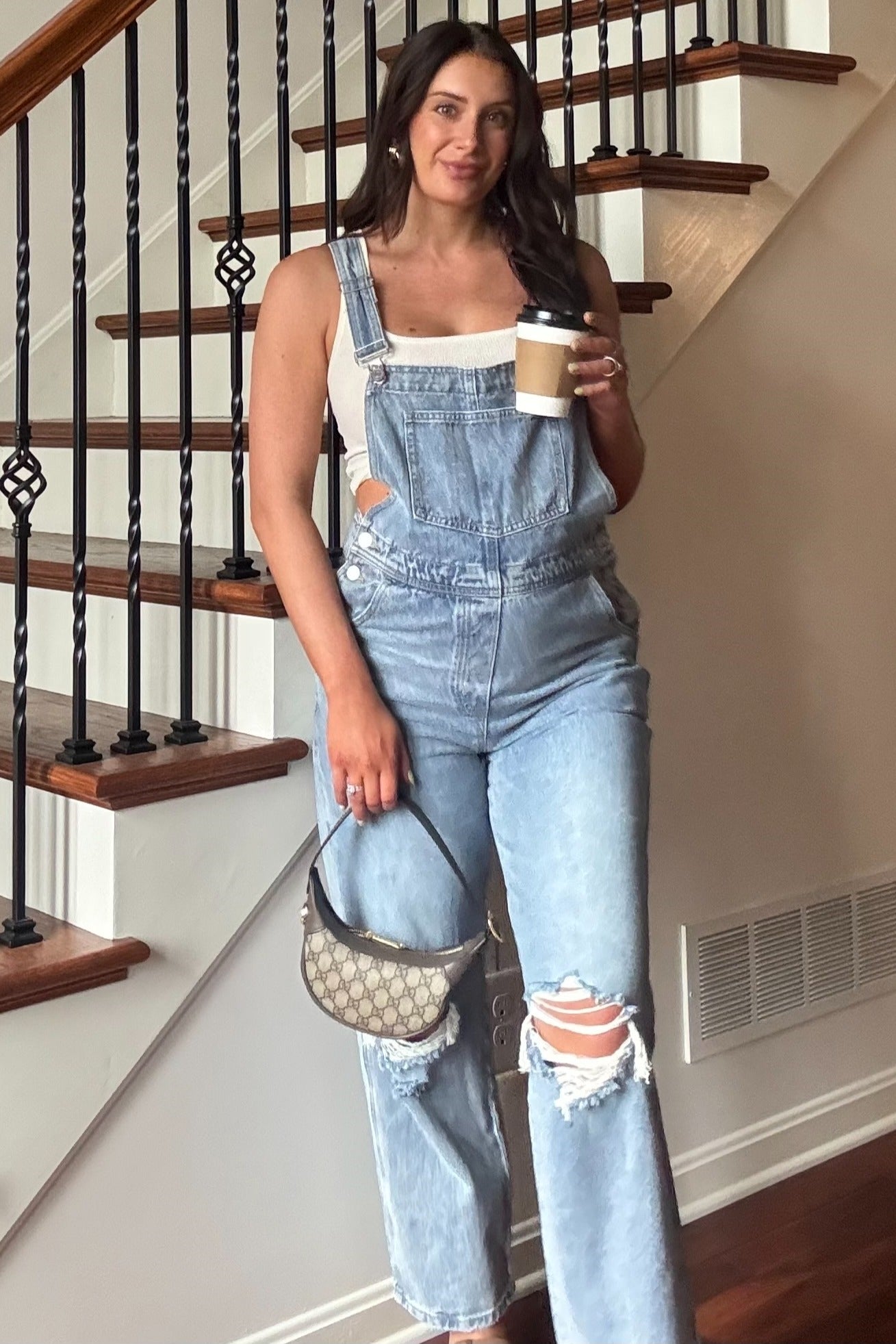 woman in light wash distressed denim overalls and white tank top holding coffee and a designer handbag by a staircase