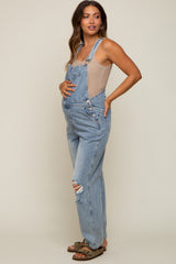 Pregnant woman wearing light wash denim maternity overalls and a beige tank top against a neutral background