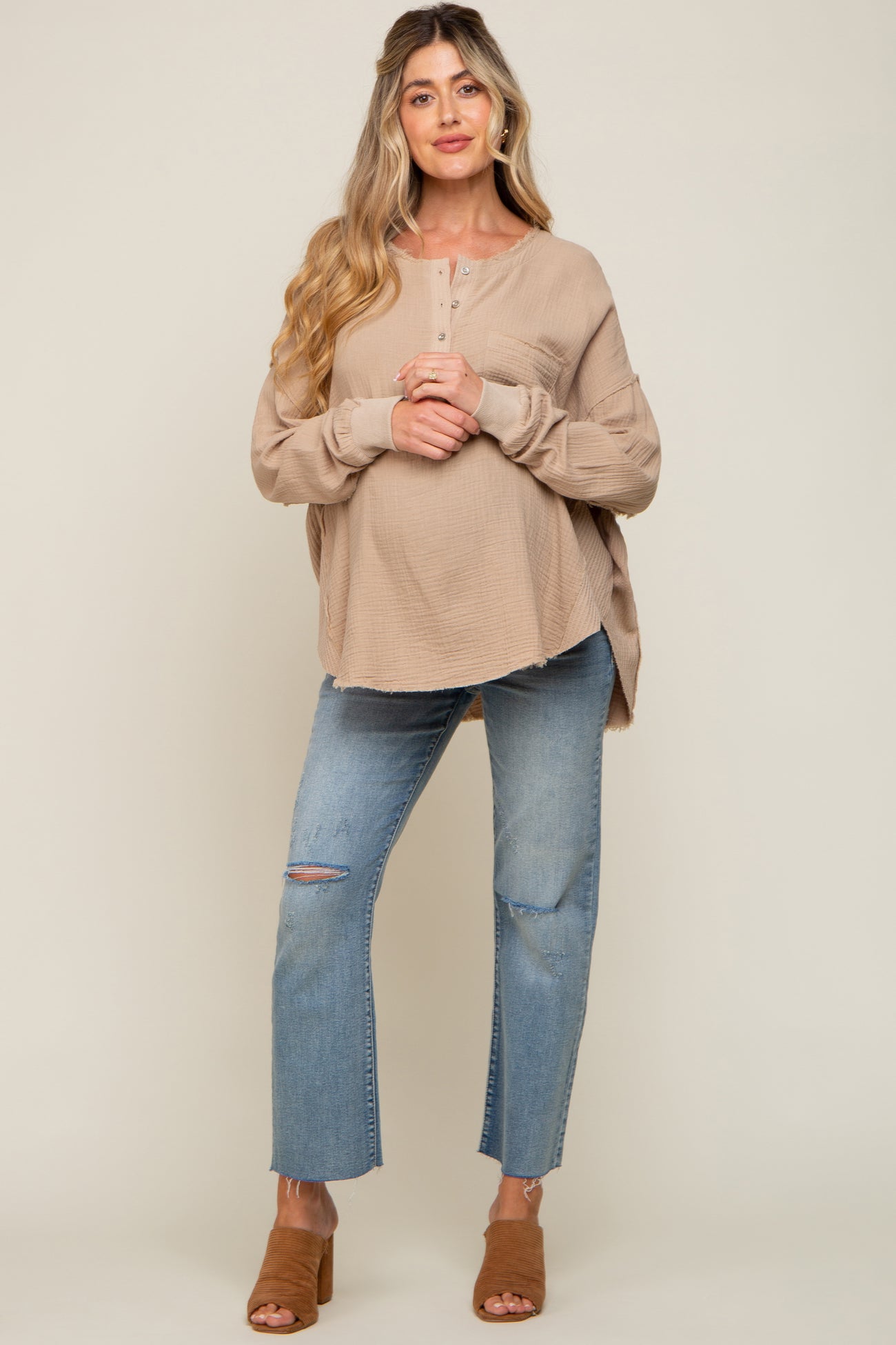 woman wearing a beige maternity top with button detailing, holding her baby bump, paired with distressed jeans and tan mules