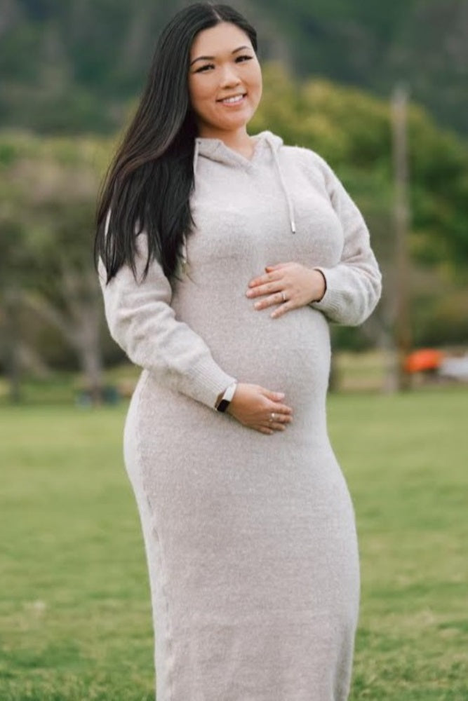 Smiling pregnant woman in a beige hooded sweater dress holding her baby bump outdoors in a park