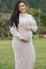 Smiling pregnant woman in a beige hooded sweater dress holding her baby bump outdoors in a park