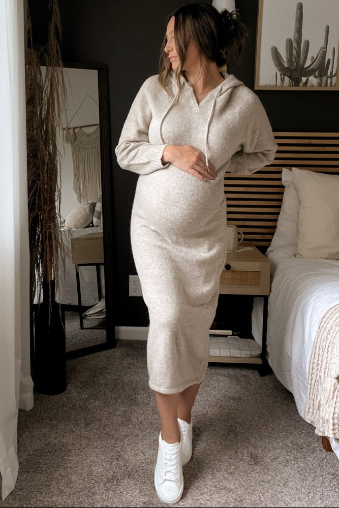 woman wearing a light beige maternity hoodie dress and holding her baby bump while standing beside a bed