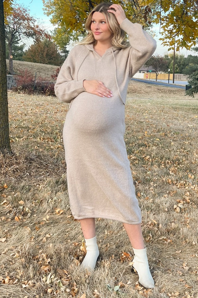 pregnant woman in a beige knit maternity hoodie dress and white ankle boots standing in a field with autumn leaves