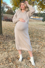 pregnant woman in a beige knit maternity hoodie dress and white ankle boots standing in a field with autumn leaves