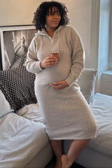 woman wearing a cozy beige maternity hooded dress, holding a drink and showing her baby bump while sitting on a couch