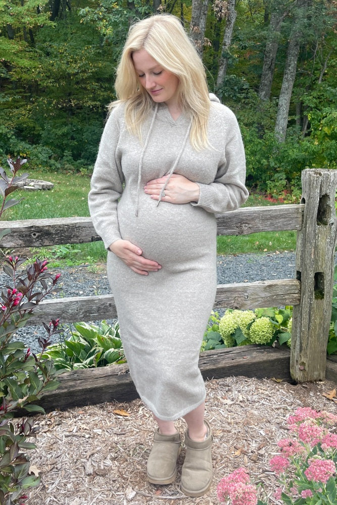 pregnant woman in beige knit hoodie dress holding her baby bump outdoors by a wooden fence