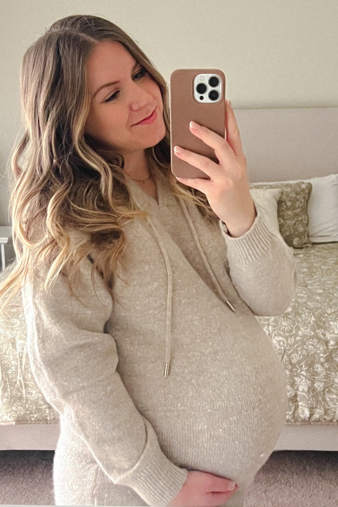 woman wearing a cream maternity sweater with a hood, showing her baby bump while taking a mirror selfie