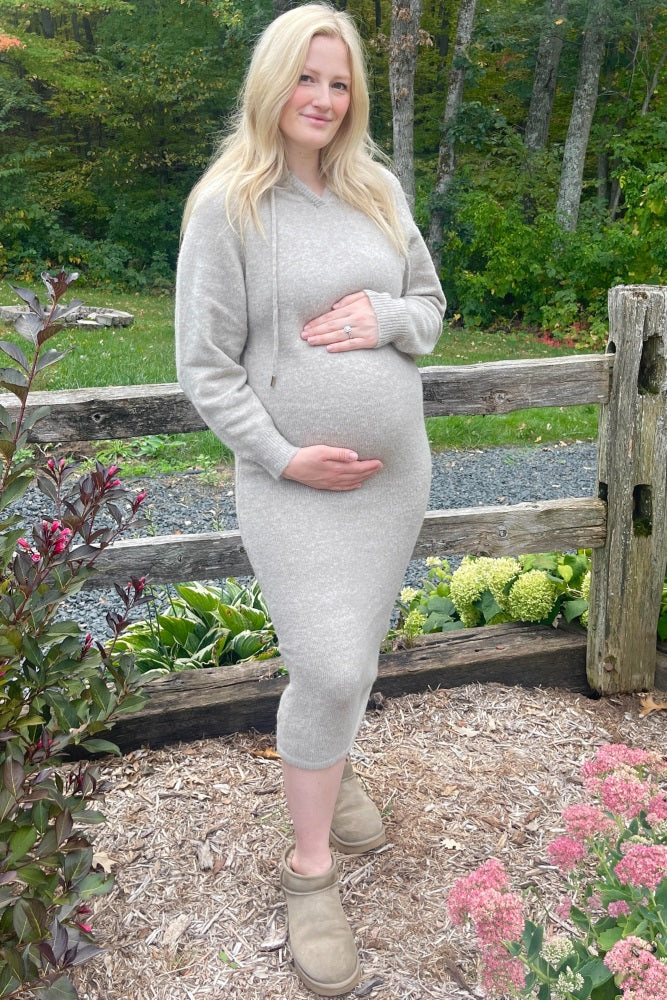 woman wearing a light grey maternity sweater dress and holding her baby bump standing outdoors