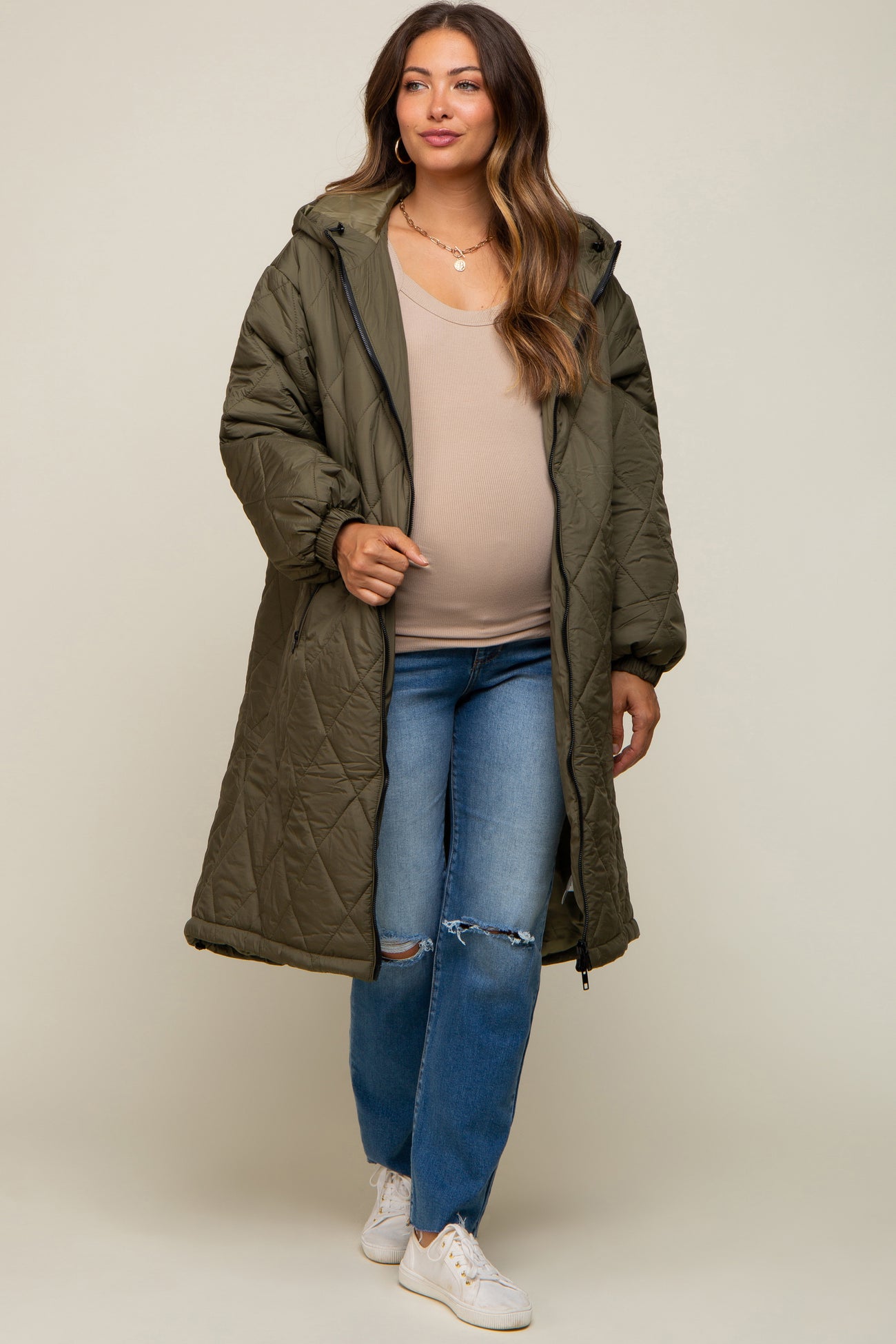 woman wearing an olive green quilted maternity coat over a beige top and blue ripped jeans, showing her baby bump