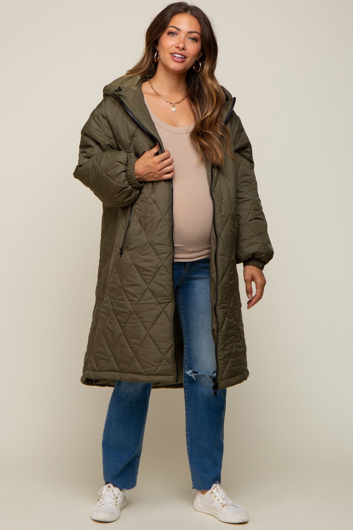 woman wearing an olive green long quilted maternity coat over a beige top, with a visible baby bump and blue jeans