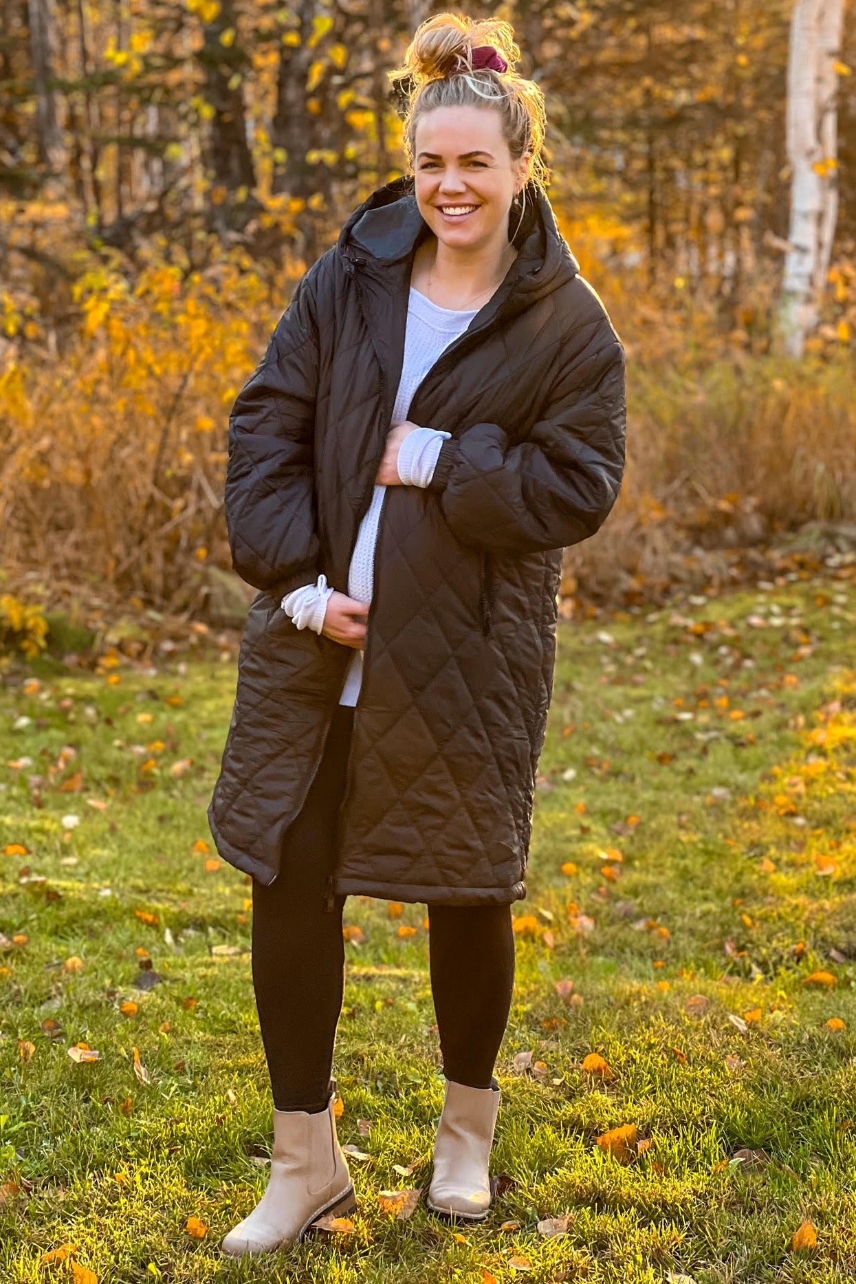 woman wearing a black quilted maternity coat and holding her baby bump in an autumn setting