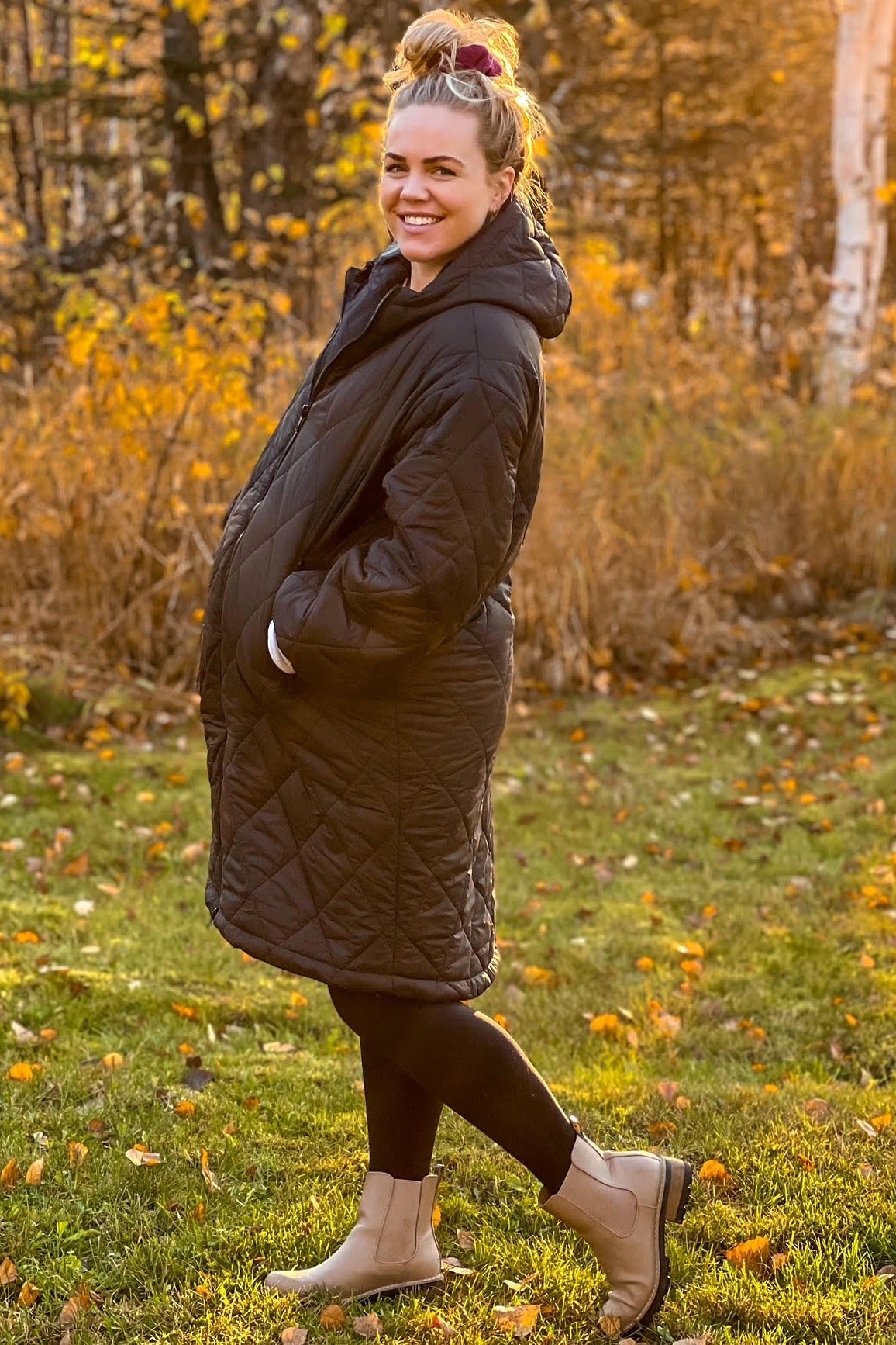 Smiling woman in a black quilted maternity coat and tan boots standing in a park with autumn leaves
