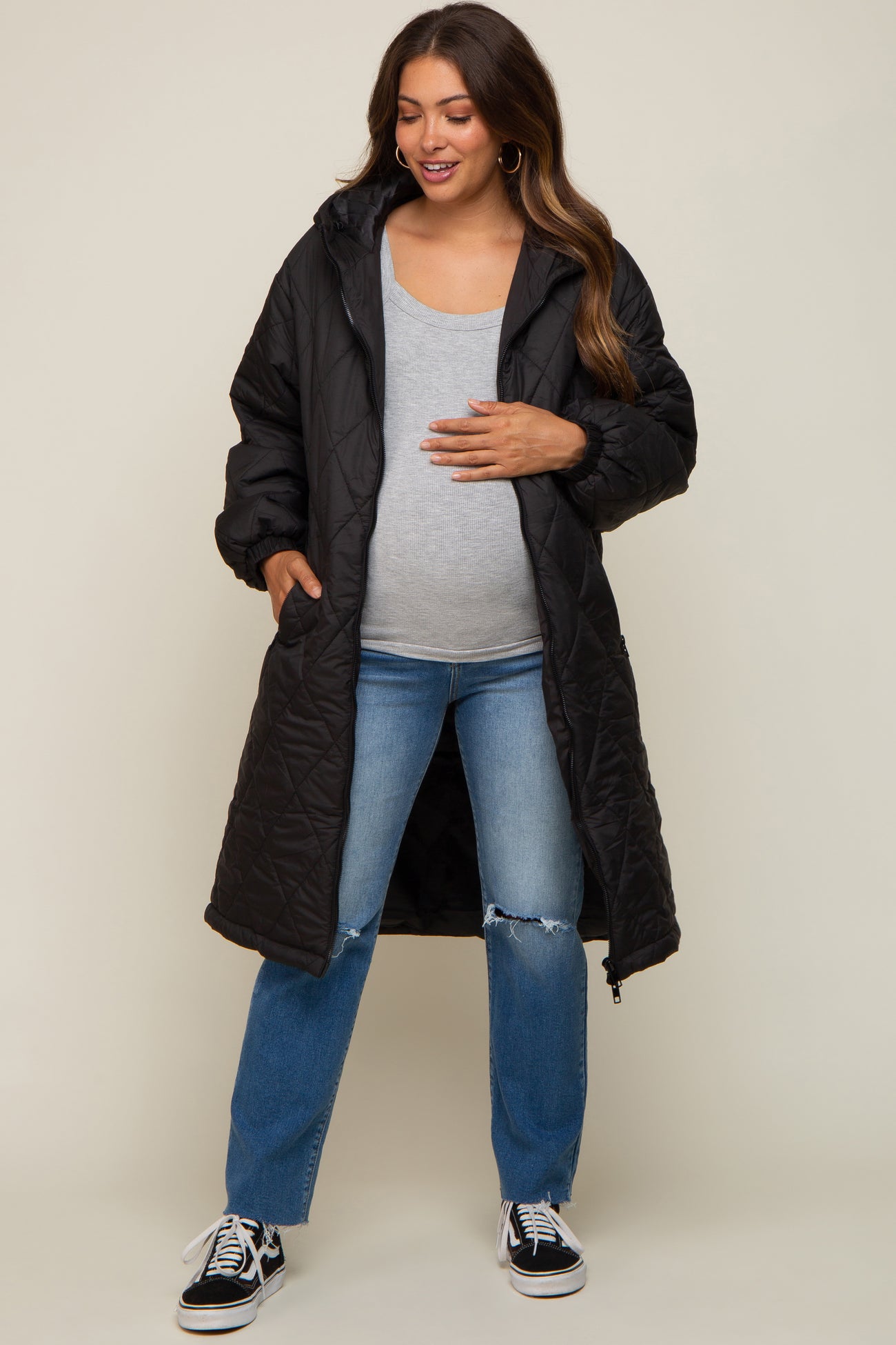 woman wearing a black long quilted maternity coat over a gray top and blue jeans, gently holding her baby bump