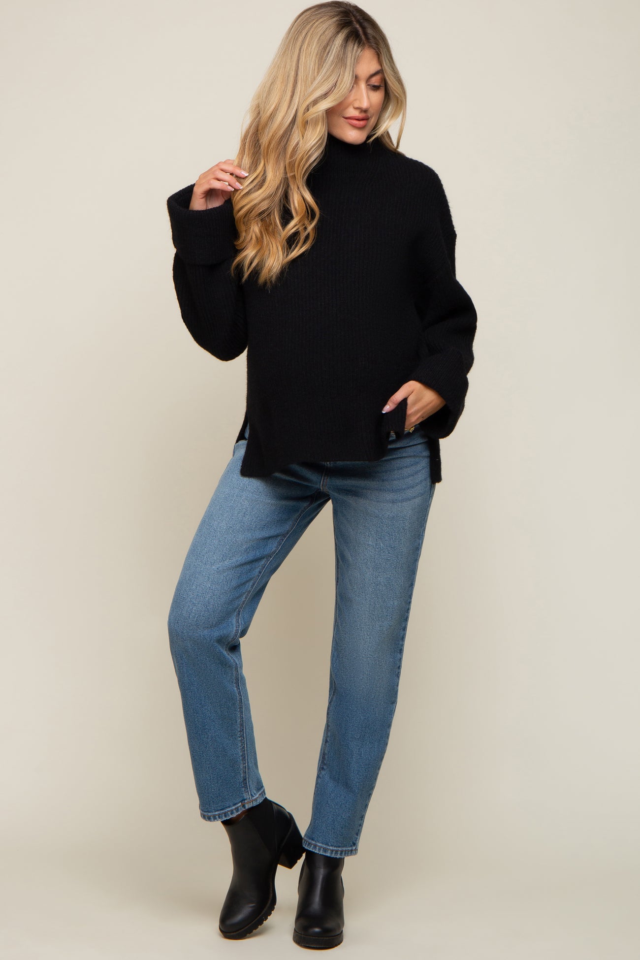 woman wearing a black oversized maternity sweater with side slits and blue straight-leg jeans, posing with one hand on her hip