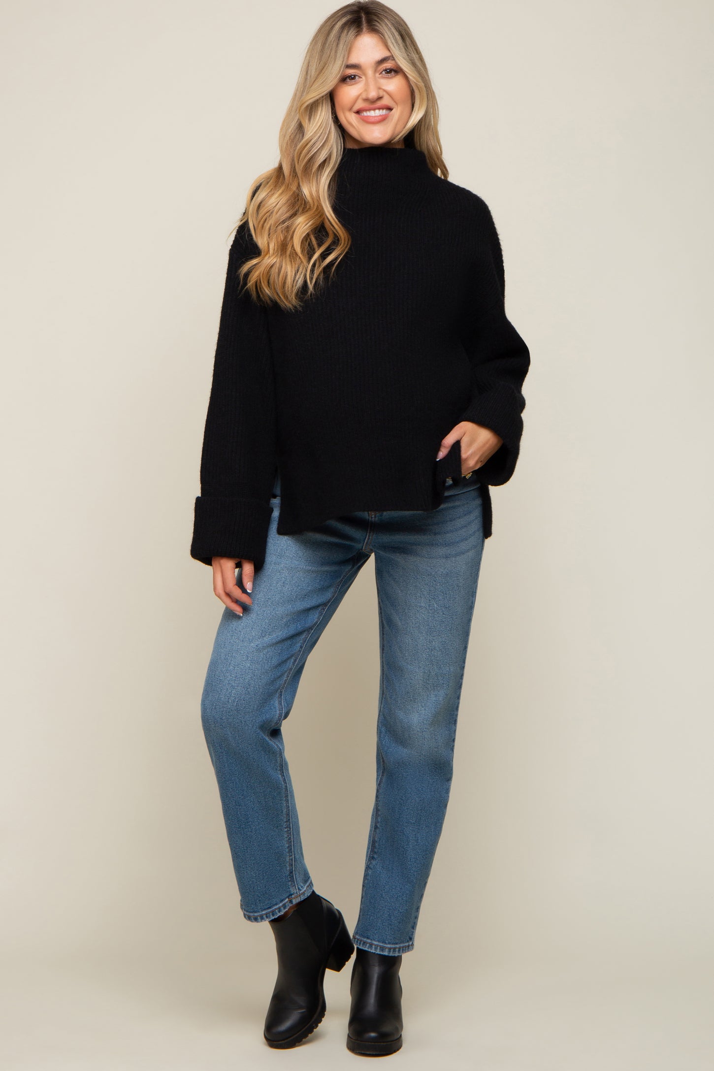 woman wearing a black oversized knit maternity sweater with wide sleeves and blue jeans, standing and smiling