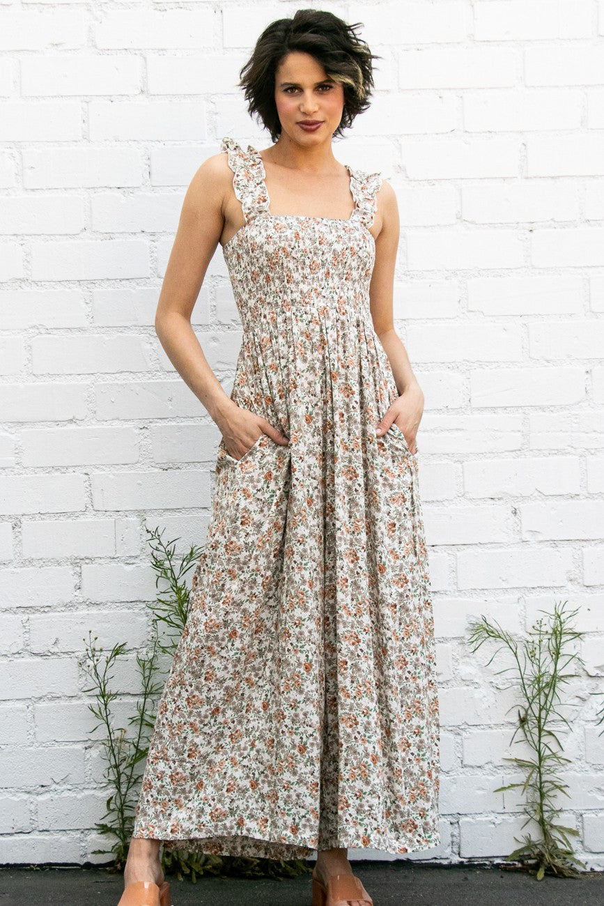 woman wearing a floral maxi dress with ruffle straps and pockets, standing against a white brick wall