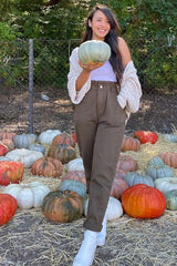 woman in brown pants and white cardigan holding a pumpkin in a pumpkin patch with various colorful gourds