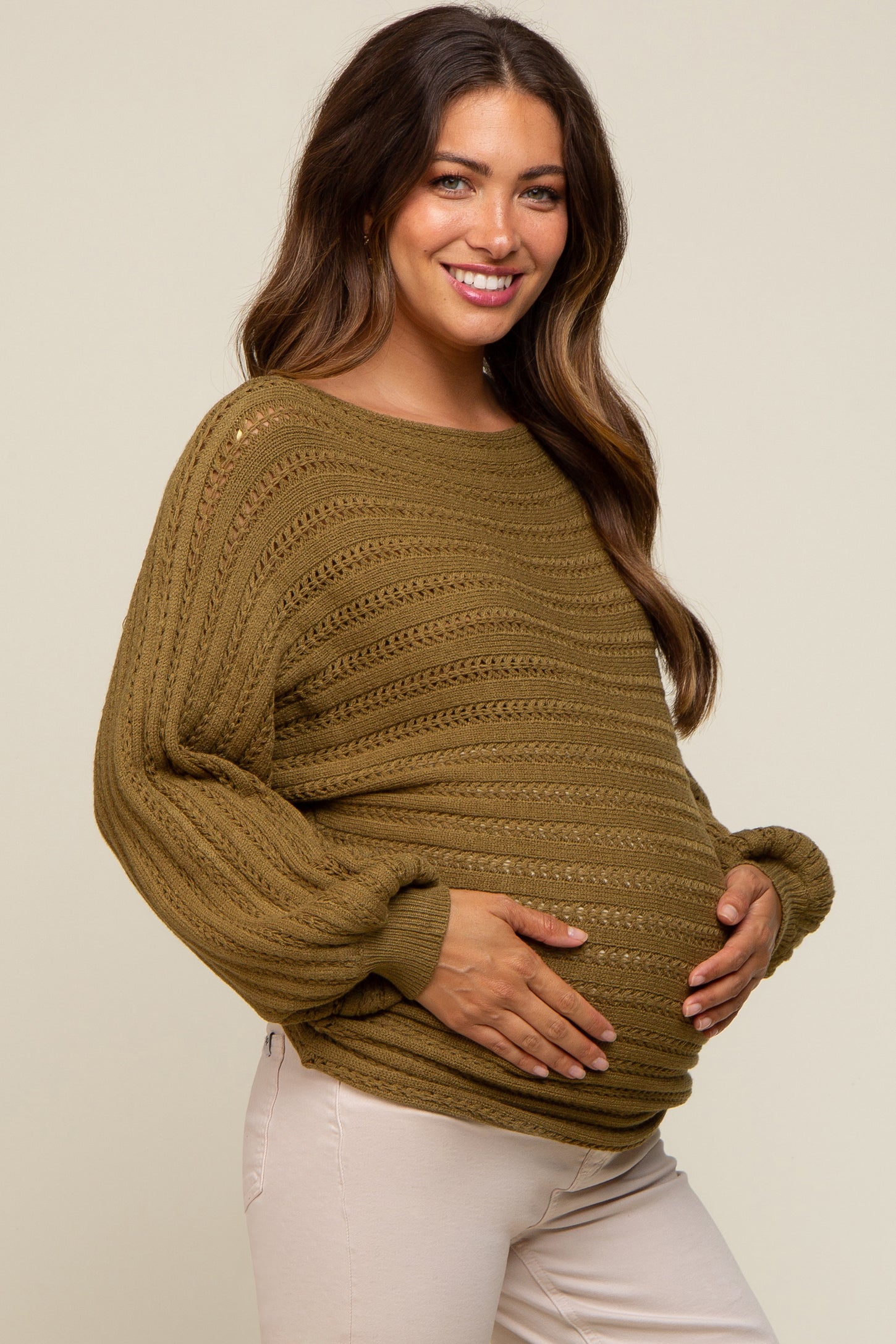 smiling pregnant woman wearing an olive green crochet knit maternity sweater and white pants