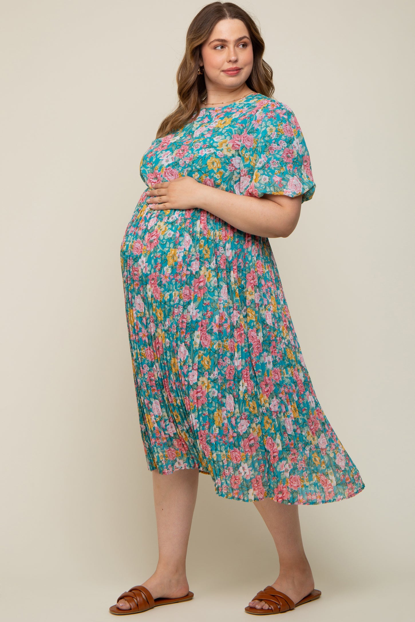 woman wearing a floral pleated maternity dress in turquoise, showcasing her baby bump, standing on a neutral background